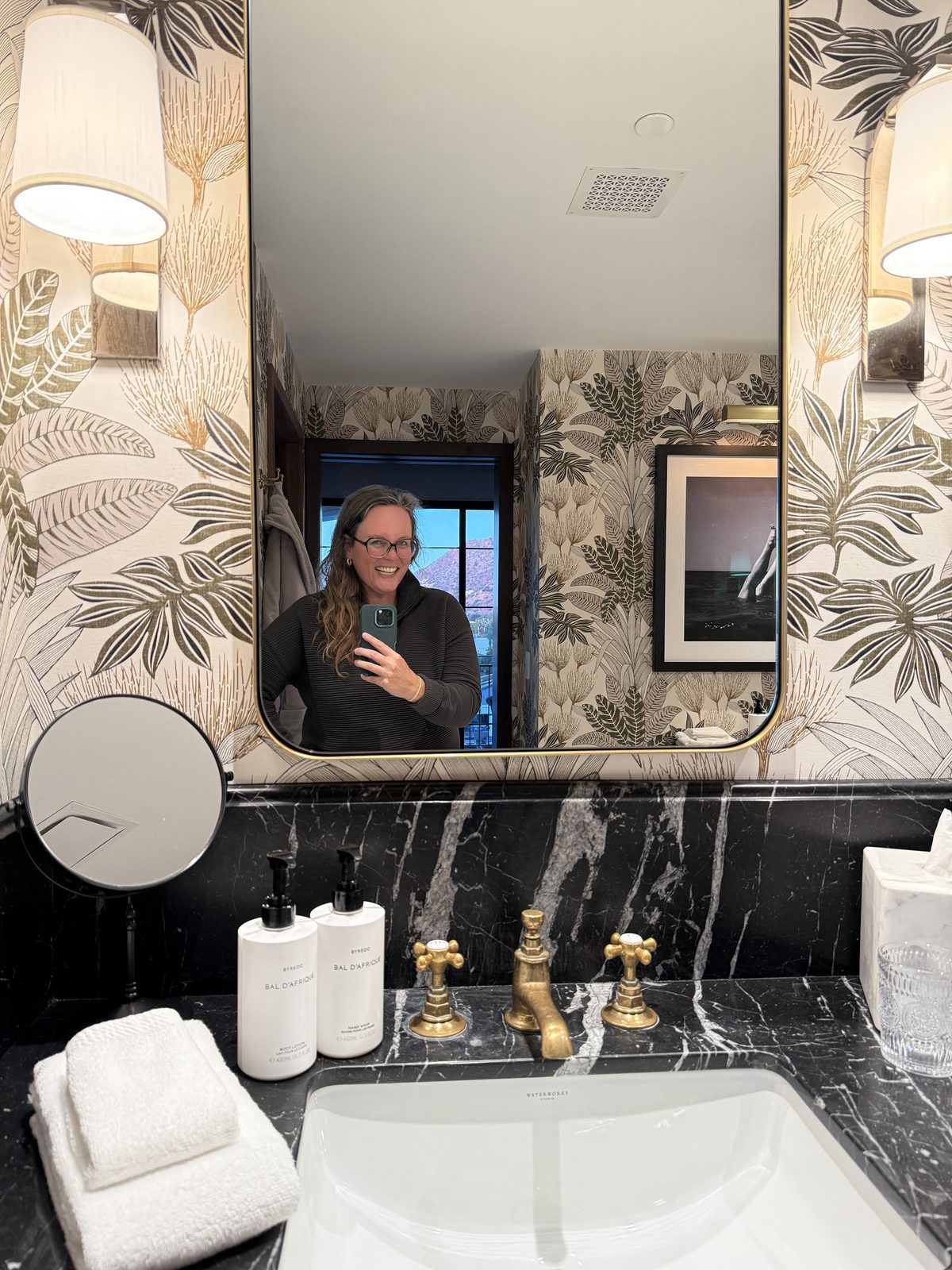 Black marble bathroom vanity with brass fixtures and Byredo toiletries in a Terrace Suite at The Global Ambassador, with Camelback Mountain visible through the window