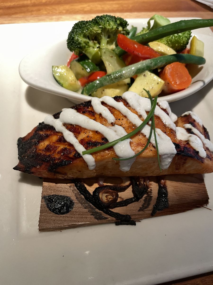 Close up of cedar plank grilled salmon with a cream sauce and a bowl of steamed vegetables.