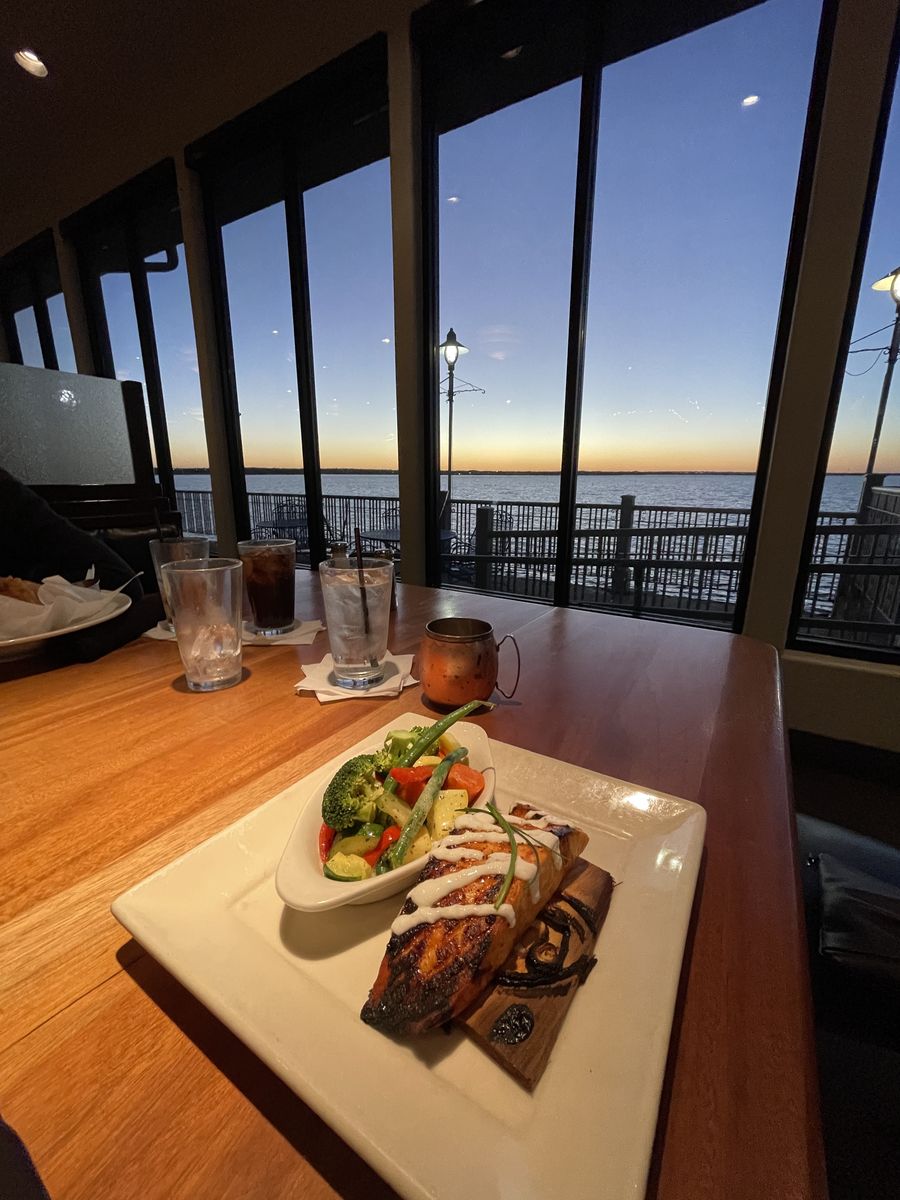 A plate of grilled salmon and steamed vegetables on a table by the window at Hefner Grill, with Lake Hefner at sunset behind it.