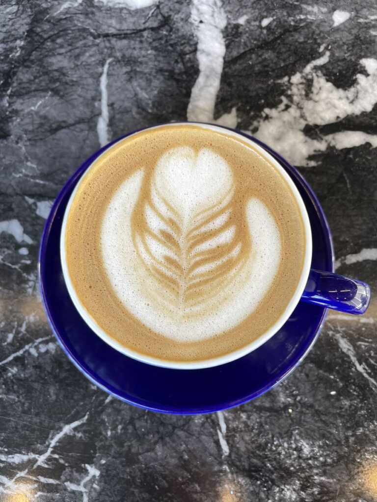 An oat milk latte with leaf latte art in a blue cup on a dark marble table at Harvey Bakery and Kitchen.