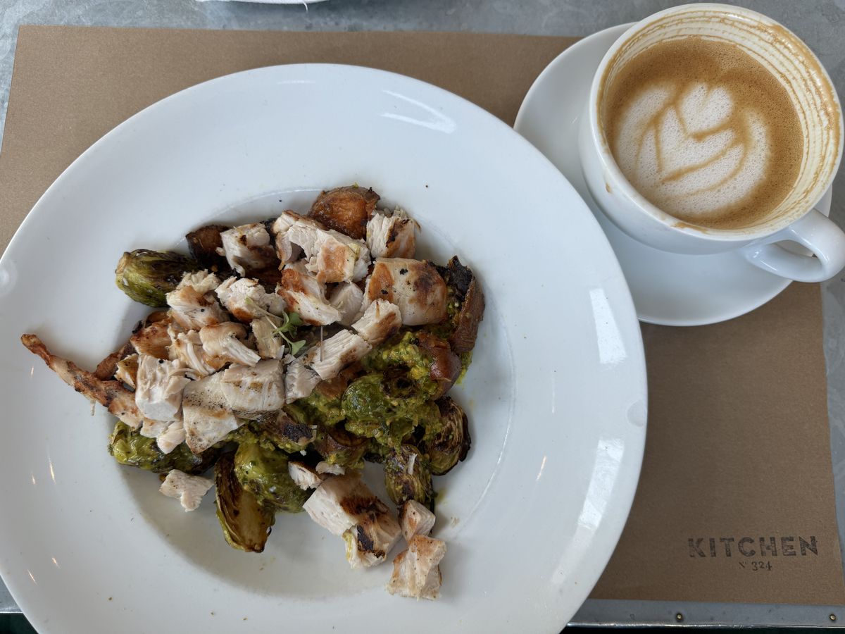 A modified breakfast bowl of roasted brussels sprouts, sweet potato and grilled chicken beside a latte on a Kitchen No. 324 tray.